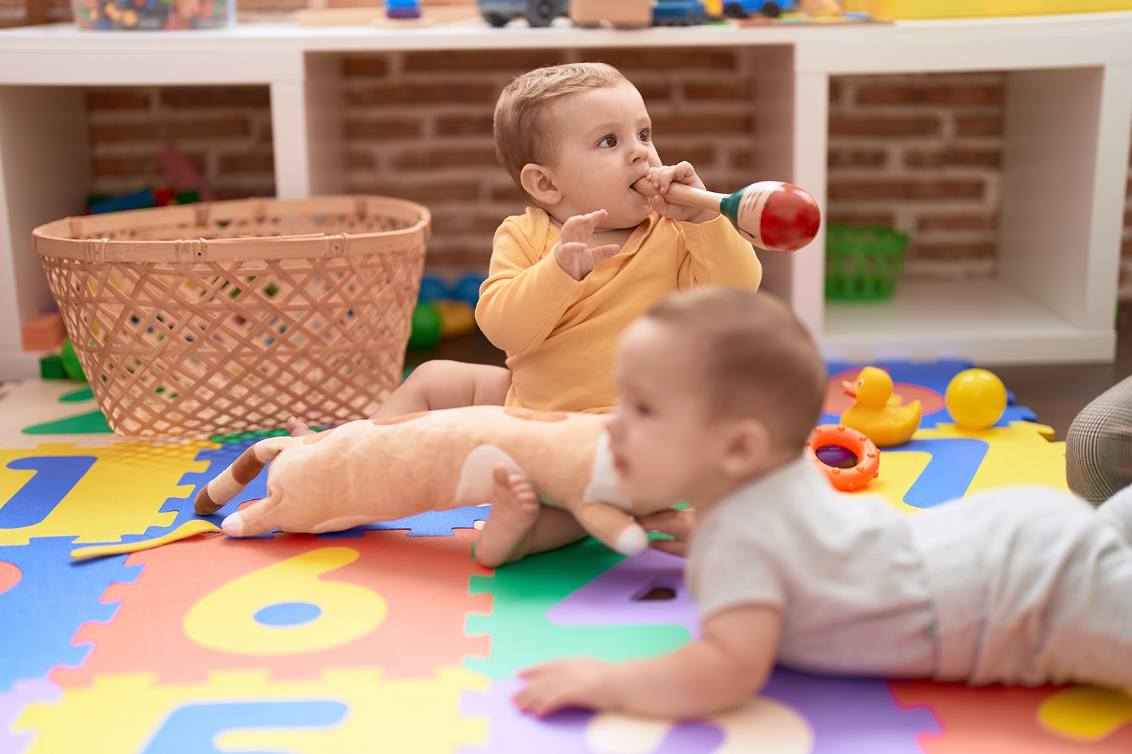 Two toddlers sucking maraca sitting on floor at kindergarten