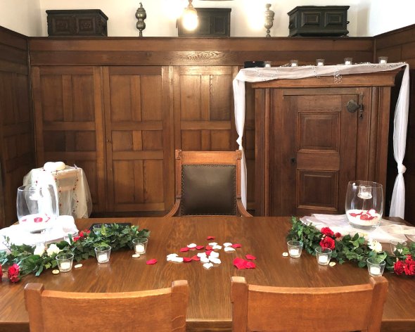 Zwei Stühle stehen vor einem hochzeitlich geschmückten Tisch, dahinter steht der Stuhl der Standesbeamten.