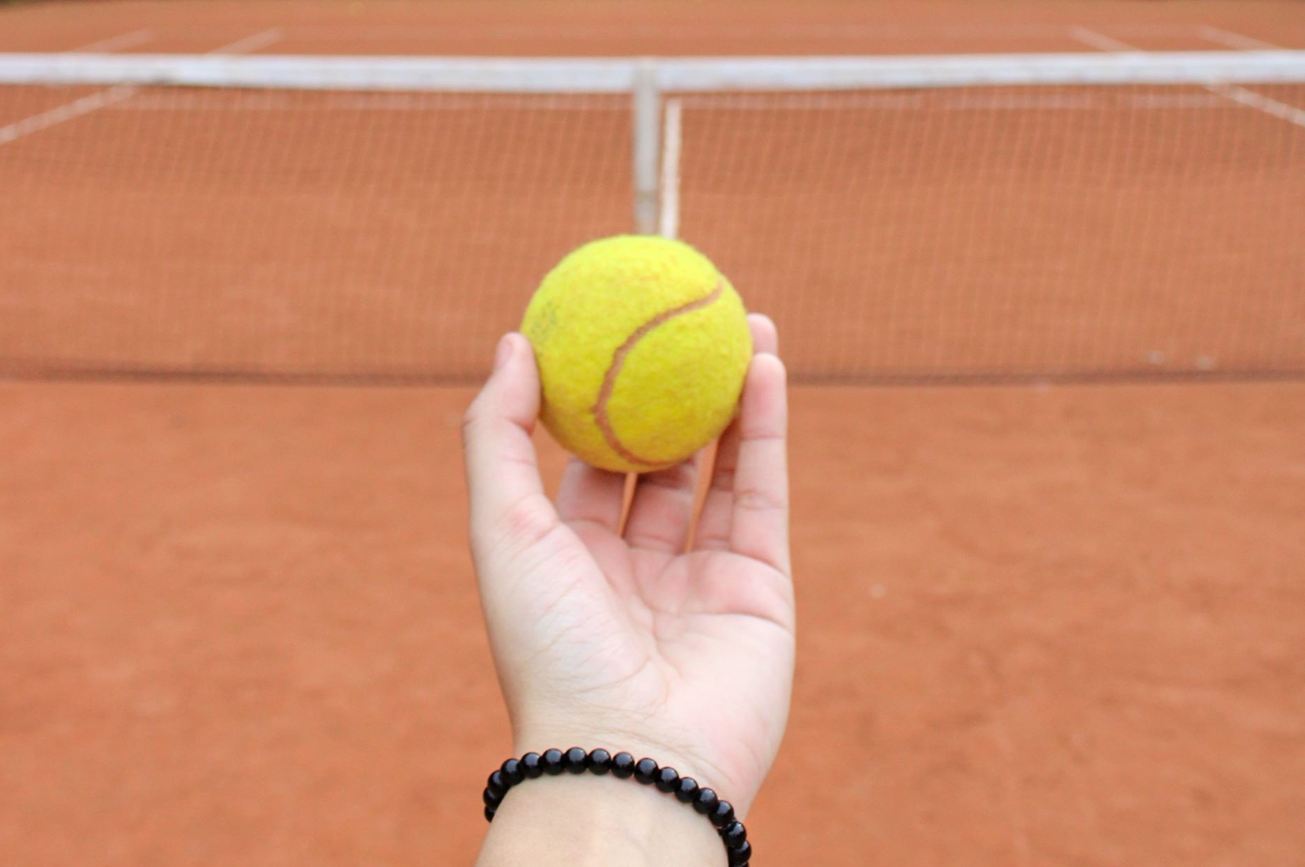 Eine Hand hält einen gelben Tennisball über einem rötlichen Tennisplatz.