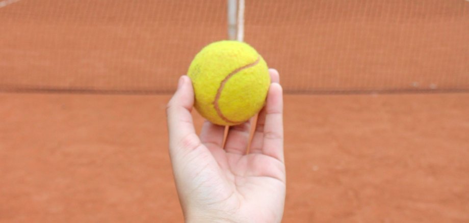 Eine Hand hält einen gelben Tennisball über einem rötlichen Tennisplatz.