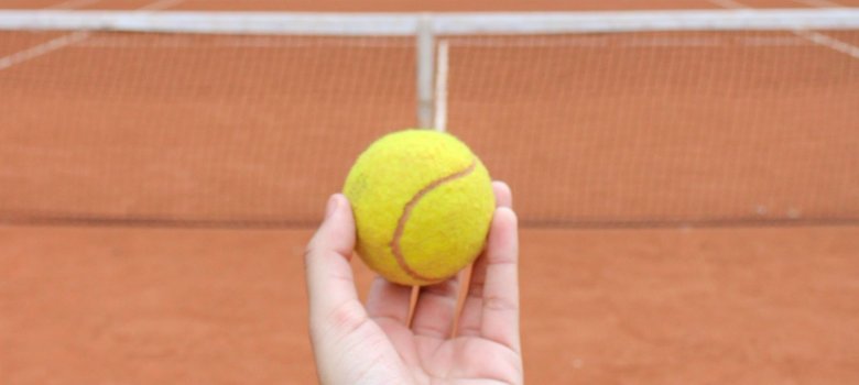 Eine Hand hält einen gelben Tennisball über einem rötlichen Tennisplatz.